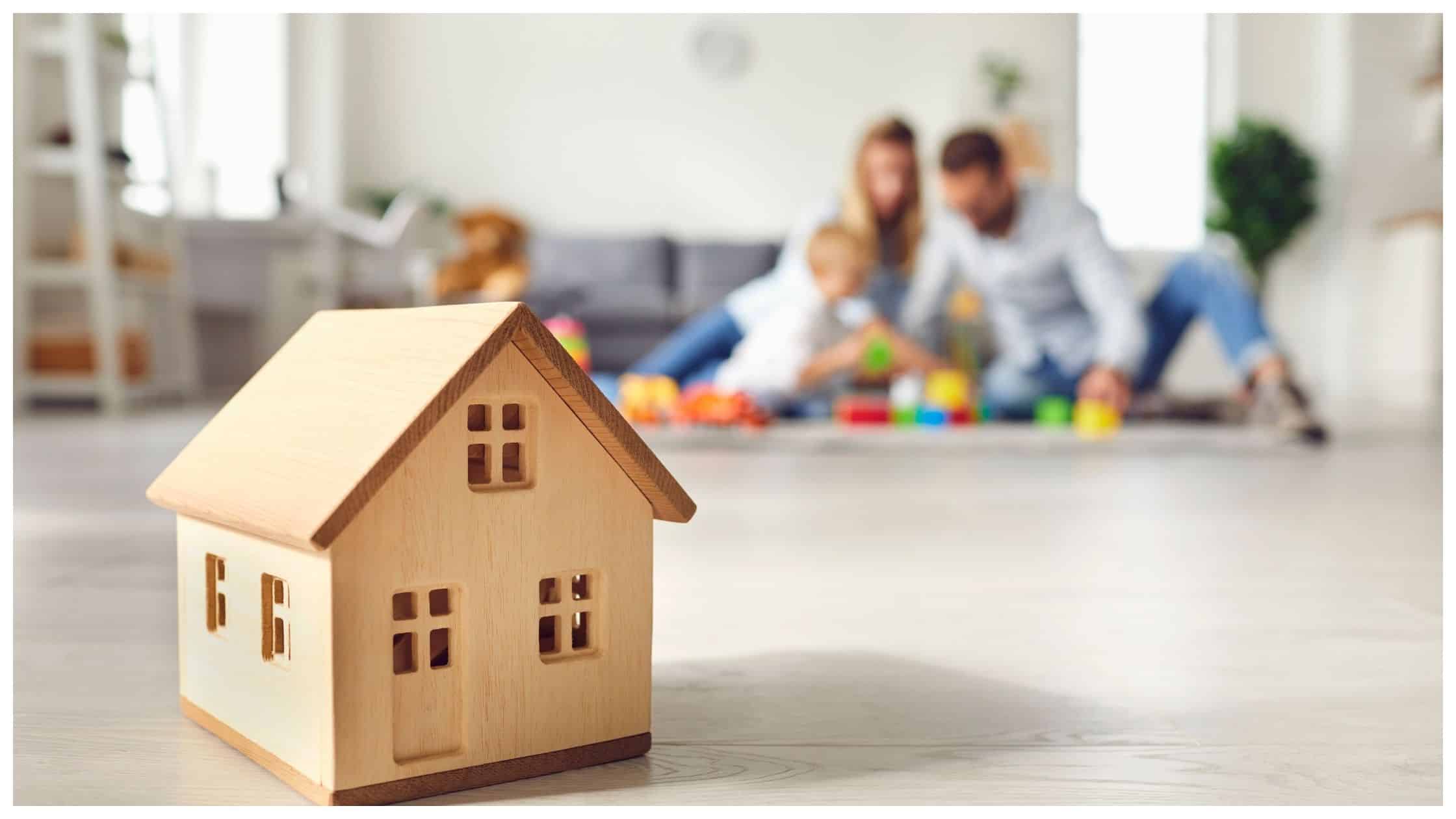 Little Wooden House on Floor of Cozy Room with Happy Family Playing in Background
